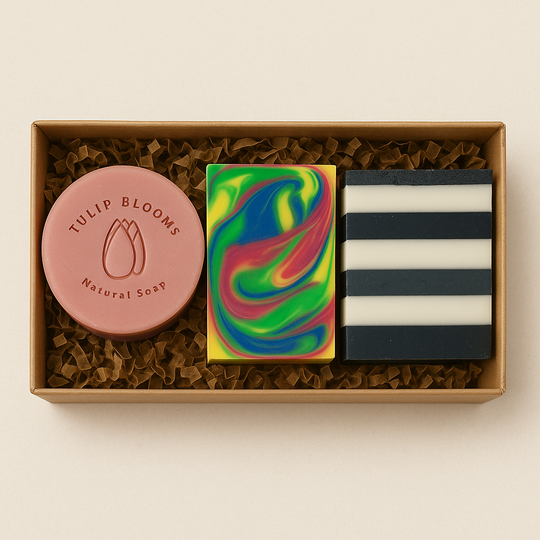 A kraft gift box filled with three Tulip Blooms handmade soaps — one pink round bar, one rainbow swirl bar, and one black-and-white striped bar, nestled in crinkle paper.