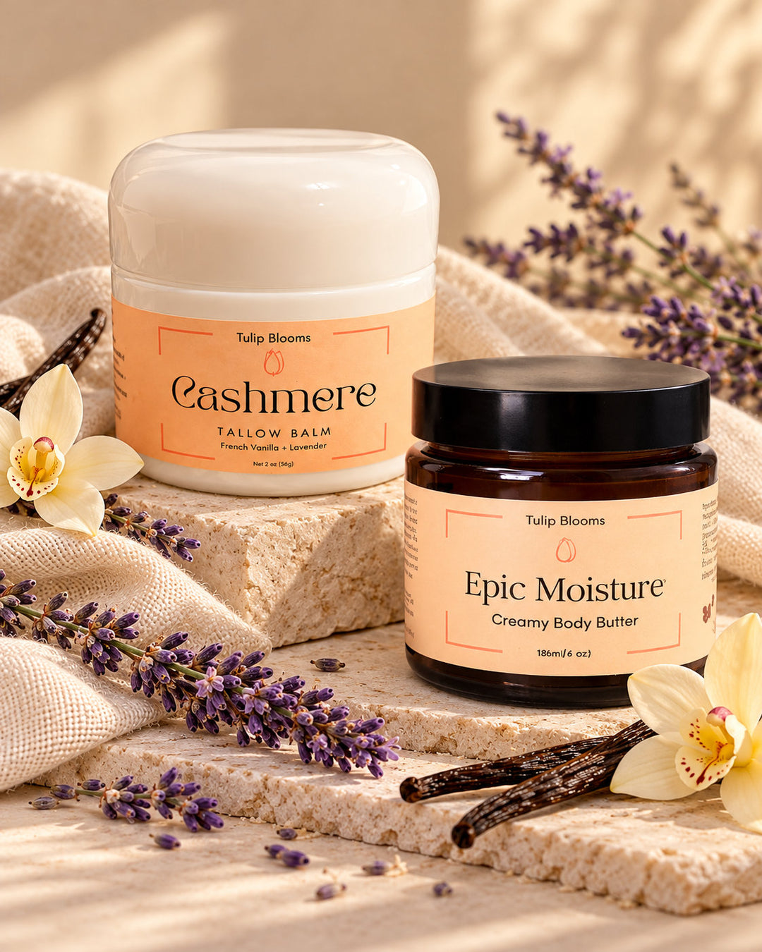 Two Tulip Blooms body care products from Tulip Dreams: 'Cashmere' Tallow Balm in a white jar and 'Epic Moisture' Creamy Body Buffer in an amber glass jar, styled on stone blocks with lavender, vanilla beans, and white orchids.