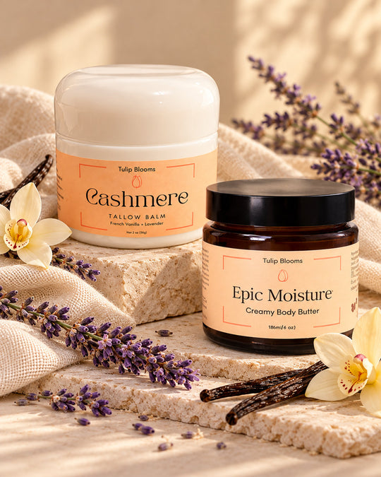 Two Tulip Blooms body care products from Tulip Dreams: 'Cashmere' Tallow Balm in a white jar and 'Epic Moisture' Creamy Body Buffer in an amber glass jar, styled on stone blocks with lavender, vanilla beans, and white orchids.