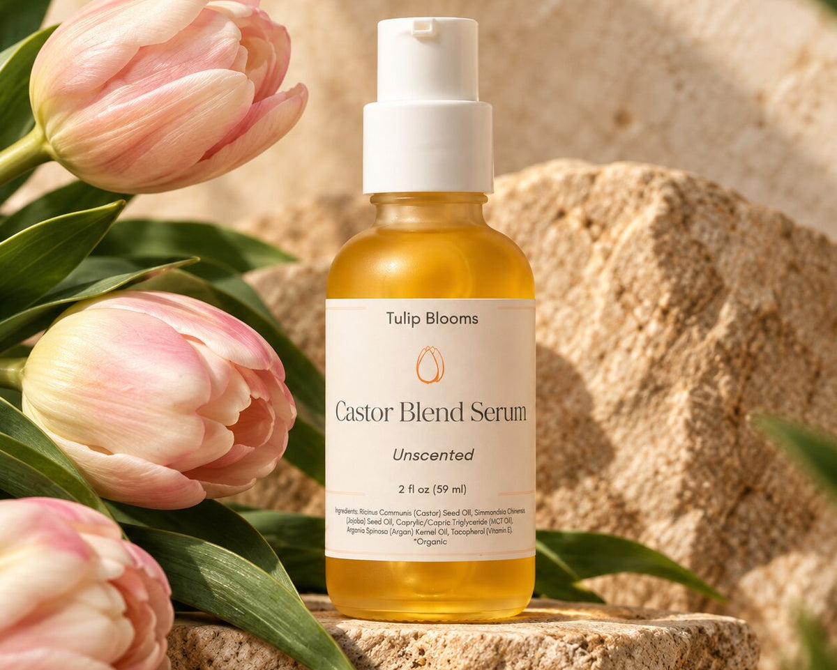 A bottle of Tulip Blooms Castor Blend Serum stands on a stone surface with pink tulips and green leaves around it. The bottle contains golden yellow liquid and has a white pump cap and cream label.